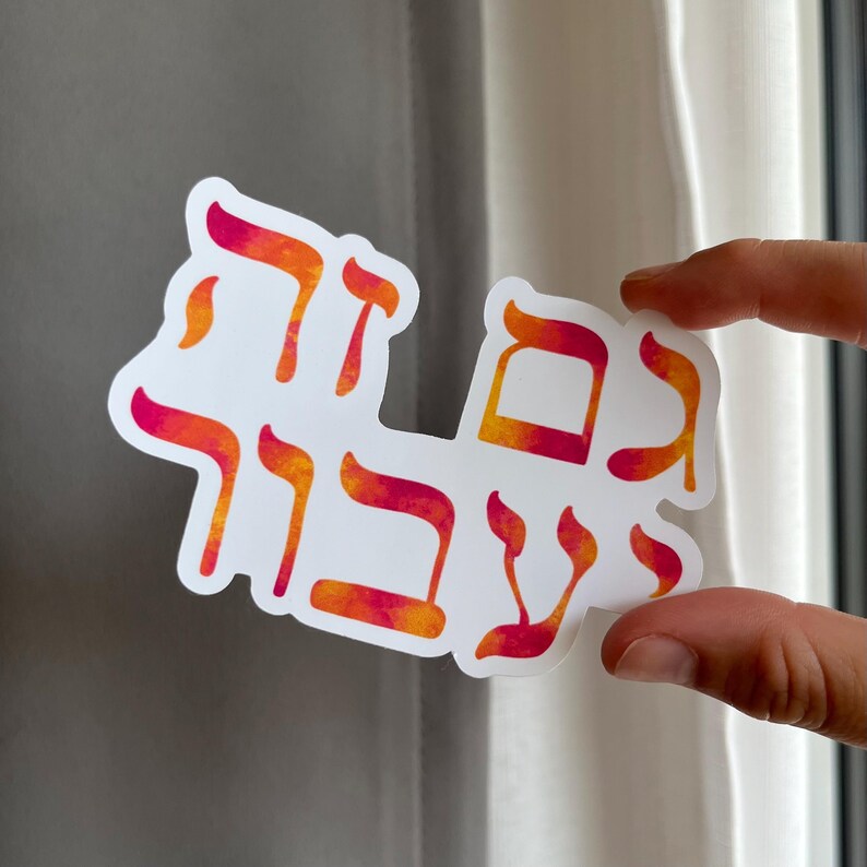 May include: White sticker with Hebrew text in orange and pink watercolor lettering. The sticker is held by a hand in front of a window. The text appears to be a phrase or saying.
