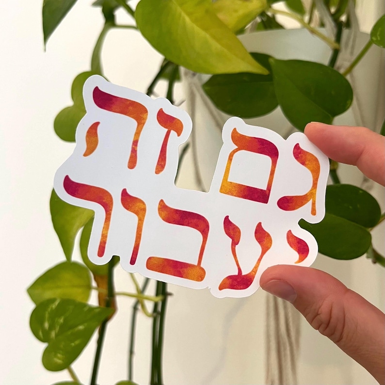 May include: A white sticker with Hebrew text in orange and red gradient colors. The sticker is held in front of green leafy plants. The text appears to be a phrase or saying.