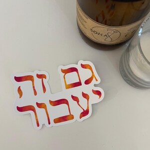 May include: A white sticker with Hebrew text in orange and red watercolor style. The sticker is on a white surface, with a brown candle and a clear glass jar in the background.