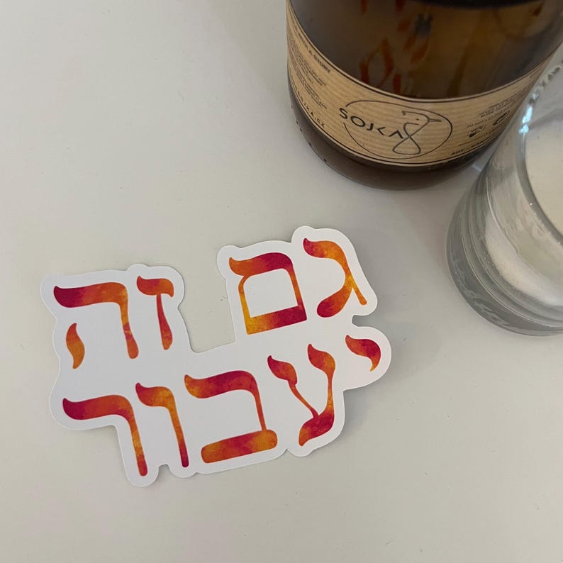 May include: A white sticker with Hebrew text in orange and red watercolor style. The sticker is on a white surface, with a brown candle and a clear glass jar in the background.