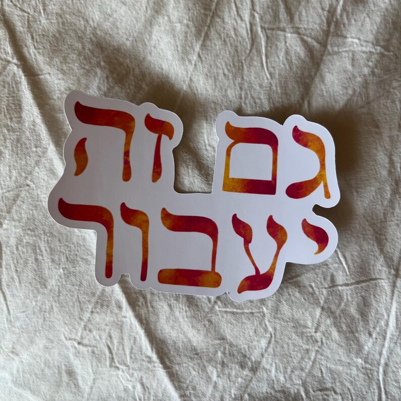 May include: A white sticker with Hebrew text in red and orange watercolor-style lettering. The sticker has a unique shape, with the text arranged to fit the contours of the words. The sticker is on a beige fabric background.