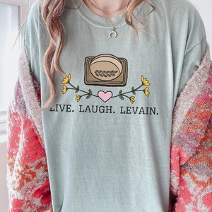 May include: A light blue t-shirt with a graphic of a loaf of bread in a brown tray with a pink heart and yellow flowers. The text "LIVE. LAUGH. LEVAIN." is printed below the graphic.