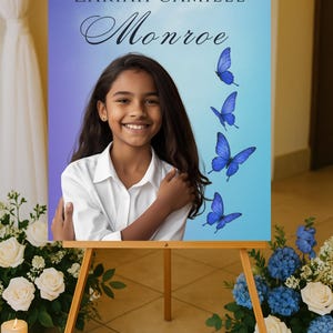 May include: A wooden easel displays a sign with the text "Celebration of Life" and the name "Zariah Camille Monroe." The sign features a photo of a smiling girl and blue butterfly graphics. Floral arrangements and candles surround the easel.