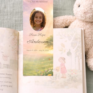 May include: A bookmark featuring a portrait of a smiling child with the text "You Are My Sunshine" and the name "Sarai Hope Anderson" with birth and death dates. The bookmark is placed in an open book with illustrations and a stuffed bunny.