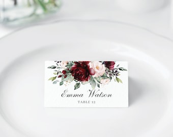 May include: A white place card with a burgundy and blush floral design. The text on the card reads "Emma Watson, Table 12".