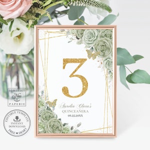 May include: A gold glitter number 3 table number card with a green floral design and gold geometric frames. The text "Aurelia Clara's Quinceañera 08.25.20XX" is at the bottom of the card.