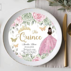 May include: A white plate with a floral design featuring pink roses and green foliage. The plate has gold text that reads "Mis Quince" and includes the name "Aurelia Clara" and the date "08.25.20XX". A gold crown and butterflies are also featured.