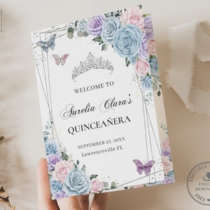May include: A folded invitation card with a floral and butterfly design. It features a silver tiara, the words "Welcome to Aurelia Clara's Quinceañera", and the date and location. The card is decorated with blue, purple, and pink roses.