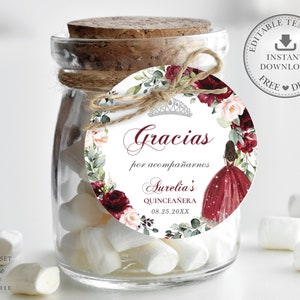 May include: A round favor tag with a watercolor floral design in shades of burgundy, pink, and green. The tag features a crown, the text "Gracias por acompa&ntilde;arnos Aurelia's Quincea&ntilde;era 08.25.20XX", and a silhouette of a girl in a burgundy dress.