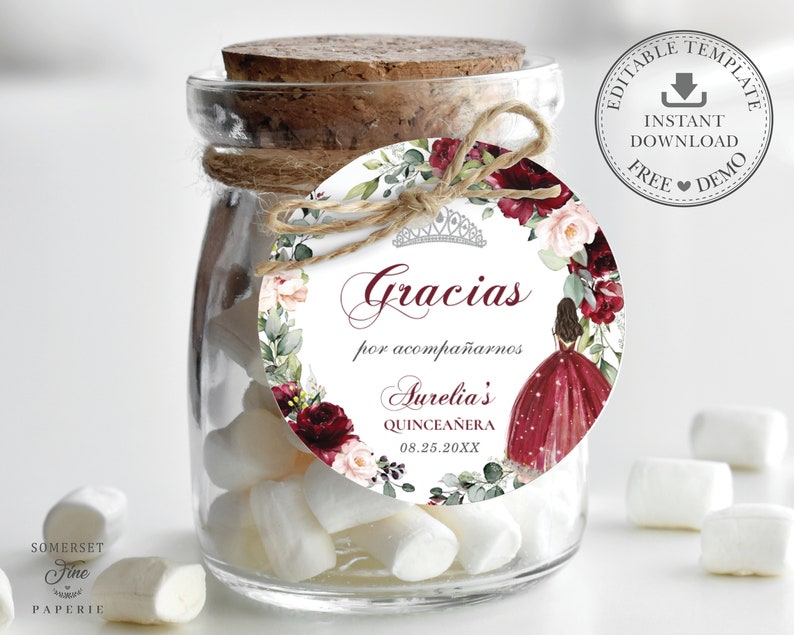 May include: A round favor tag with a watercolor floral design in shades of burgundy, pink, and green. The tag features a crown, the text "Gracias por acompa&ntilde;arnos Aurelia's Quincea&ntilde;era 08.25.20XX", and a silhouette of a girl in a burgundy dress.