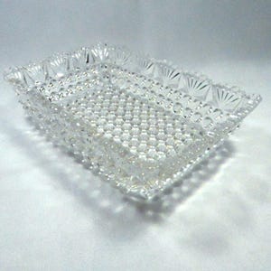 May include: A rectangular, clear glass dish with a textured, bubble-like pattern on the base and sides. The rim features a decorative, fan-like design. The dish is likely vintage and suitable for serving or display.