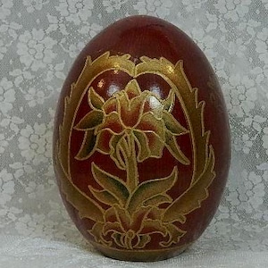May include: A decorative, egg-shaped object with a deep red finish. The surface features a gold-colored floral design. The egg is standing upright on a white surface, with a floral patterned background.