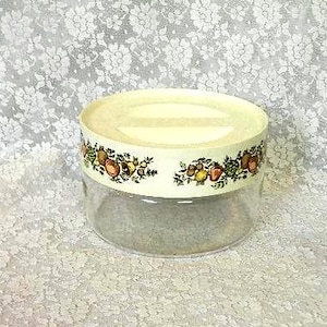 May include: A clear glass jar with a cream-colored lid. The lid is decorated with a band of colorful fruit and foliage. The jar is sitting on a white lace surface.