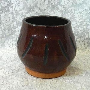 May include: A handmade, brown ceramic cup with a glossy finish. The cup features vertical, dark brown, carved lines and a natural, unglazed base. The interior is dark brown. The cup is likely used for drinking beverages.