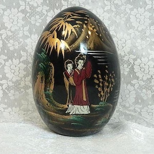 May include: Black, egg-shaped decorative item with a painted scene of two figures in traditional attire. The artwork features gold accents, green foliage, and a landscape design. The egg is displayed on a white surface.