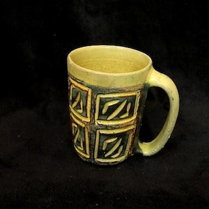 May include: A ceramic mug with a pale yellow rim and handle, featuring a textured body decorated with a grid of dark green squares. Each square contains carved, light-colored abstract markings, creating a distinct contrast against the dark background.