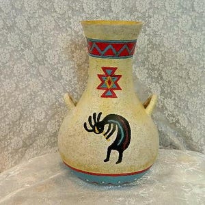 May include: A cream-colored vase with a Native American design. The vase features a Kokopelli figure, geometric patterns, and a blue and red color scheme. It has two small handles and measures approximately 10 inches tall.