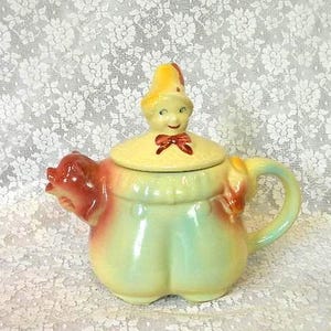 May include: A vintage ceramic teapot with a whimsical design. The teapot features a yellow lid with a face and a red bow tie. The body is a light green with red and yellow accents. The spout is shaped like a pig's head.