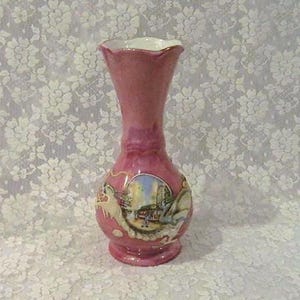 May include: A small, pink ceramic vase with a ruffled white rim. The vase features a painted scene with buildings and figures. The vase is approximately 6 inches tall.