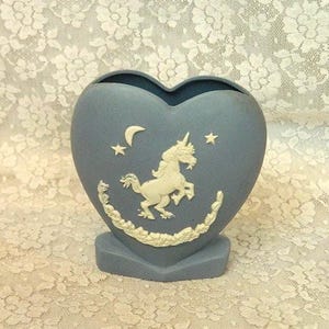 May include: A heart-shaped matte blue vase featuring a raised white relief design of a unicorn, a crescent moon, and stars. The base of the vase is also heart-shaped, and the unicorn is framed by a decorative garland of leaves and flowers.