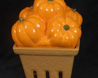 May include: A ceramic pumpkin-shaped container filled with several orange pumpkins. The pumpkins have green stems and are arranged in a tan basket-like base. The container is set against a black background.