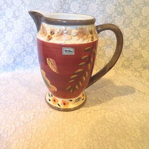 May include: A decorative ceramic pitcher with a wide base and a handle. The pitcher is primarily burgundy with a cream-colored rim and handle. It features painted leaves and floral accents, with a small label on the front.