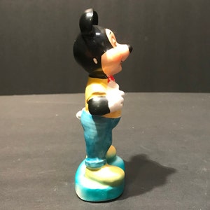 May include: A ceramic figurine of Mickey Mouse wearing a yellow shirt, blue jeans, and a red bow tie. He is standing with his arms at his sides and his head tilted to the side.