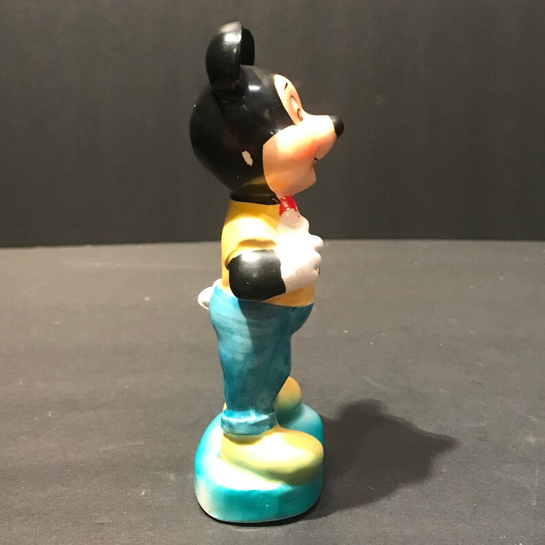 May include: A ceramic figurine of Mickey Mouse wearing a yellow shirt, blue jeans, and a red bow tie. He is standing with his arms at his sides and his head tilted to the side.