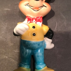 May include: A ceramic figurine of Mickey Mouse wearing a yellow shirt, blue pants, and a red bow tie. He is smiling and has his right hand on his hip.