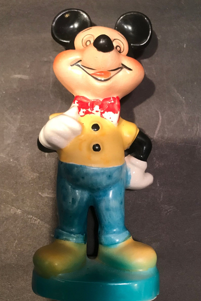 May include: A ceramic figurine of Mickey Mouse wearing a yellow shirt, blue pants, and a red bow tie. He is smiling and has his right hand on his hip.