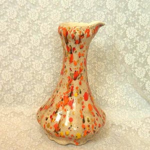 May include: A ceramic vase with a flared rim and a wide base, featuring a cream-colored background decorated with a vibrant splatter pattern of bright orange, yellow, and dark brown paint spots across the entire exterior surface of the vessel.