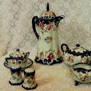 May include: A vintage porcelain tea set with a floral design. The set includes a tall coffee pot, a teapot, a sugar bowl, a creamer, and two salt and pepper shakers. The pieces are white with pink, blue, and green floral accents and dark blue trim.