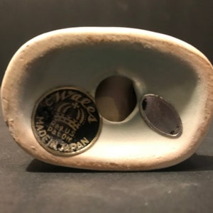 May include: Close-up of the bottom of a ceramic figurine. The figurine has a blue glaze and a black and gold sticker that reads "Wales Reg. U.S. Pat. Off. Made in Japan".