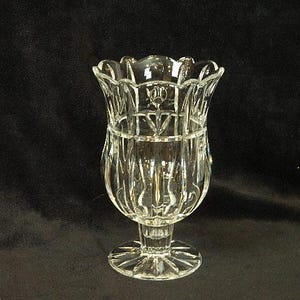 May include: A clear, cut-glass vase with a scalloped rim and a pedestal base. The vase has a detailed, textured design and is likely used for decorative purposes. The base is approximately 3 inches in diameter.