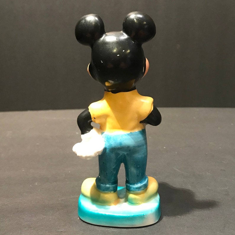 May include: A ceramic figurine of Mickey Mouse standing with his arms crossed. He is wearing a yellow shirt, blue pants, and white gloves. The figurine is standing on a blue base.