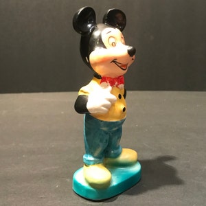 May include: A ceramic figurine of Mickey Mouse wearing a yellow shirt with a red bow tie and blue pants. He is standing on a blue base and has a friendly expression on his face.