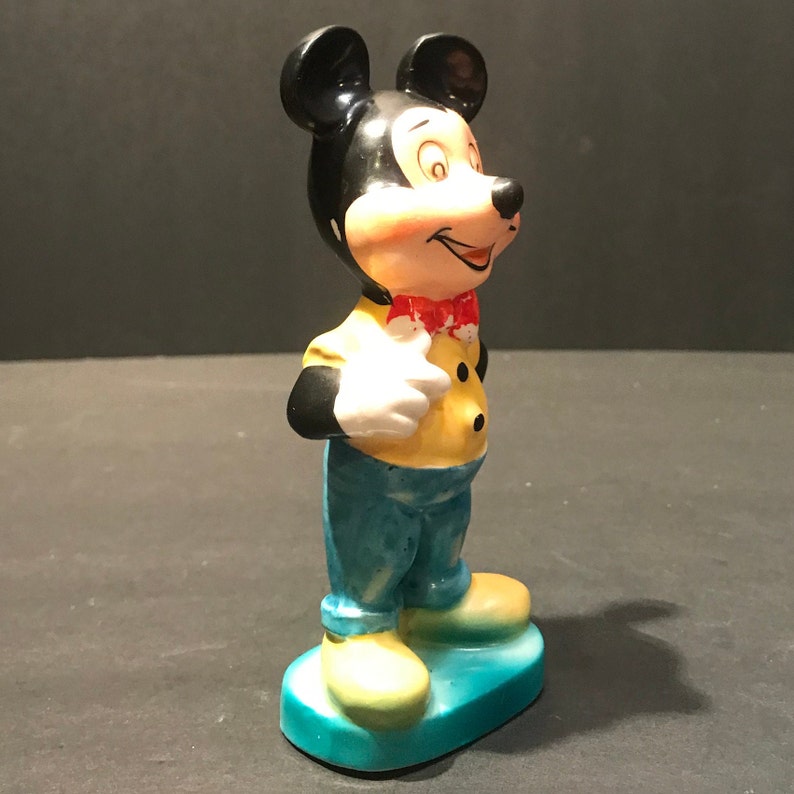 May include: A ceramic figurine of Mickey Mouse wearing a yellow shirt with a red bow tie and blue pants. He is standing on a blue base and has a friendly expression on his face.