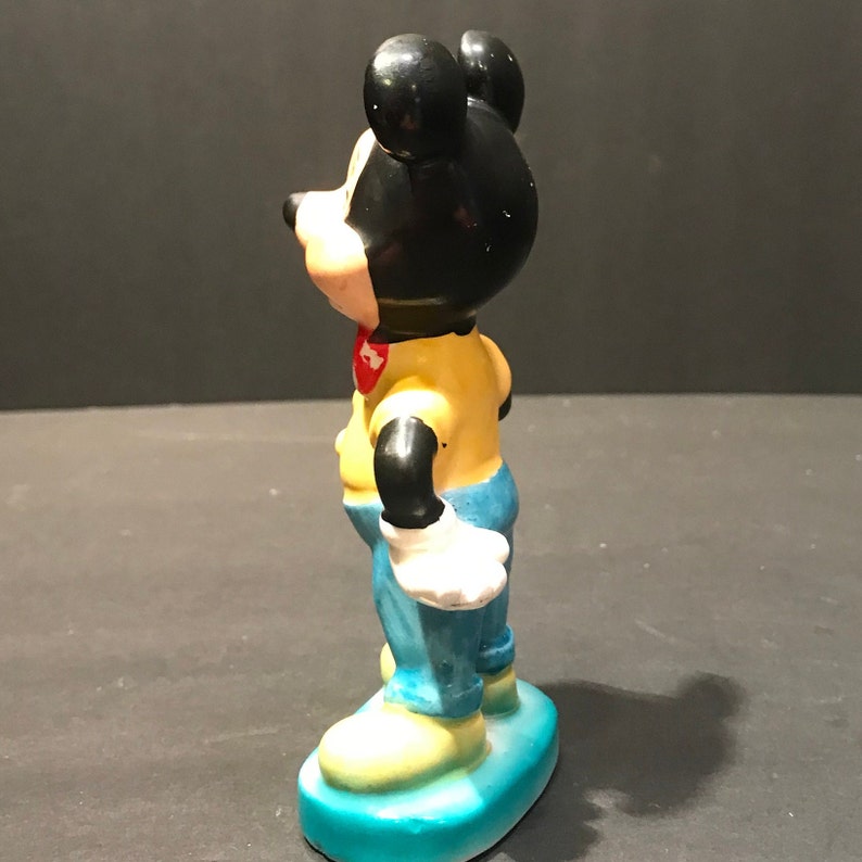 May include: A ceramic figurine of Mickey Mouse wearing a yellow shirt, blue jeans, and white gloves. He is standing on a blue base.