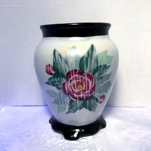 May include: White ceramic vase with a black rim and base. The vase features a hand-painted floral design with red flowers, green leaves, and gray accents. The vase has a rounded shape and a glossy finish.
