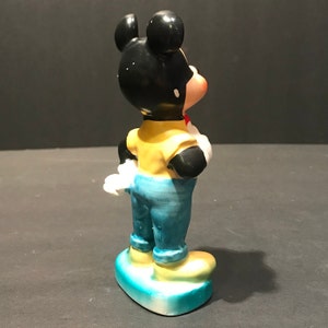 May include: A ceramic figurine of Mickey Mouse wearing a yellow shirt, blue jeans, and red shoes. He is standing on a heart-shaped base with blue and yellow paint.