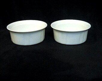 May include: Two white ceramic ramekins with a ribbed exterior. The ramekins are round with a slightly flared rim. They are set against a dark background, highlighting their clean, simple design. Ideal for baking and serving individual portions.