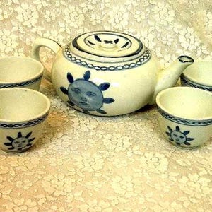 May include: A ceramic tea set consisting of a round teapot with a lid and four matching cups. The off-white pieces feature hand-painted blue sun motifs with faces and a decorative chain-link pattern around the rims and the teapot lid.