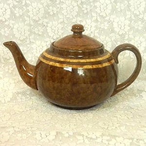 May include: A round ceramic teapot with a glossy brown mottled glaze and a horizontal band of lighter tan color around the upper body. The teapot features a curved spout, a matching lid with a small knob handle, and a curved side handle.