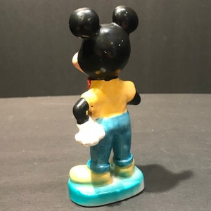 May include: A ceramic figurine of Mickey Mouse wearing a yellow shirt, blue jeans, and white gloves. He is standing with his right hand on his hip and his left hand in his pocket. The figurine is painted in bright colors and has a blue base.