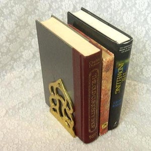 May include: A set of three hardcover books standing upright, supported by a decorative brass bookend. The books include The Old Curiosity Shop by Charles Dickens with a maroon spine and a black book titled The Indwelling with gold lettering.