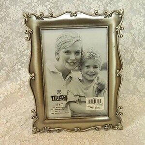 May include: A decorative gold-colored picture frame with an ornate, scrolling design. The frame holds a black and white photograph of a woman and a child. The frame is labeled "5 x 7".