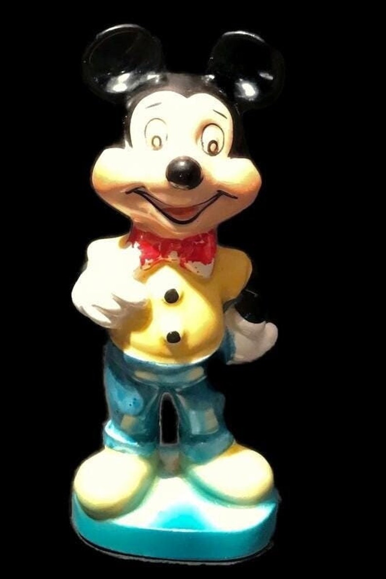 May include: A ceramic figurine of Mickey Mouse wearing a yellow shirt, blue pants, and a red bow tie. The figurine is standing on a blue base.