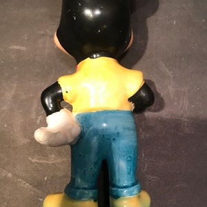 May include: A ceramic figurine of Mickey Mouse wearing a yellow shirt, blue jeans, and white gloves. The figurine is standing with its back to the camera.