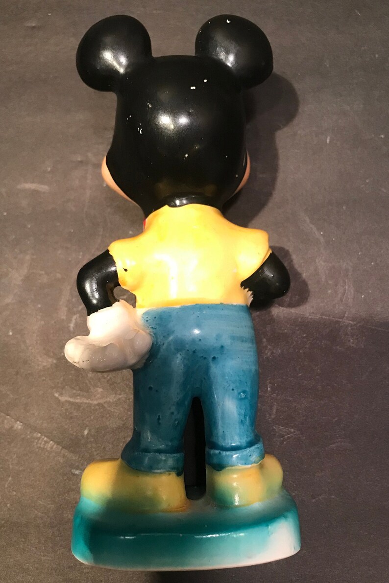 May include: A ceramic figurine of Mickey Mouse wearing a yellow shirt, blue jeans, and white gloves. The figurine is standing with its back to the camera.