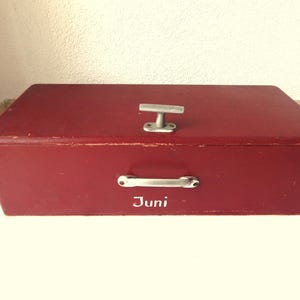 Può includere: Una scatola di legno rossa con una maniglia argentata e la parola "Juni" scritta sul davanti.